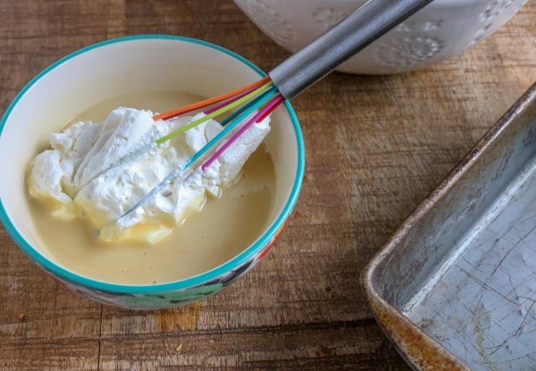 a bowl of sweetened condensed milk and whipped cream with a multi-colored whisk in it next to a white bowl and a metal pan on a brown table