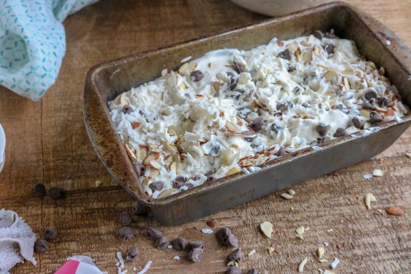 homemade ice cream topped with sliced almonds, chocolate chips and coconut in a metal pan on a brown table