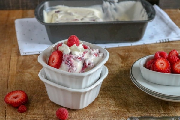 berry ice cream in a white bowl next to a white dish with berries in it on white plates with a metal pan with more ice cream in it in the background on a white cloth on a brown table