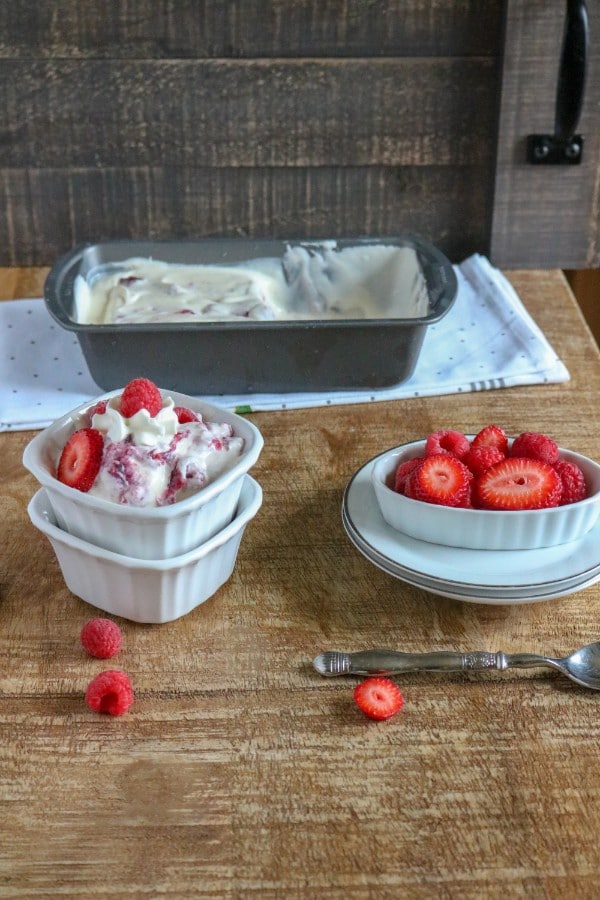 berry ice cream in a white bowl stacked on another white bowl next to berries in a white dish on white stacked plates on a brown table with a metal pan of more ice cream on a white cloth in the background