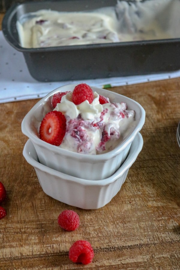 berry ice cream in a white bowl stacked on another white bowl next to berries on a brown table with a metal pan of more ice cream on a white cloth in the background