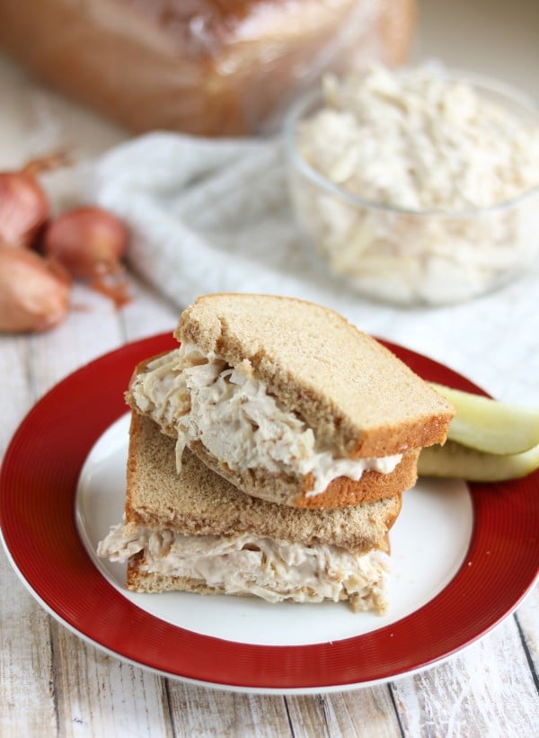 a caesar chicken salad sandwich on a red and white plate with two pickles on it on a wood table with a glass bowl of more chicken salad in it and shallots in the background