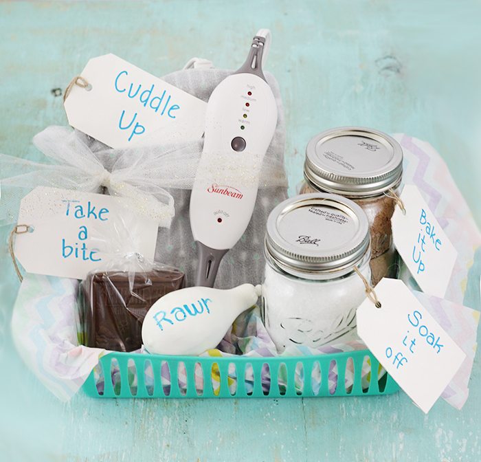 a stress relief college care package in a green plastic tray with paper tags attached to plastic bags and glass jars reading cuddle up, take a bite, bake it up, and soak it off , all on a wood table