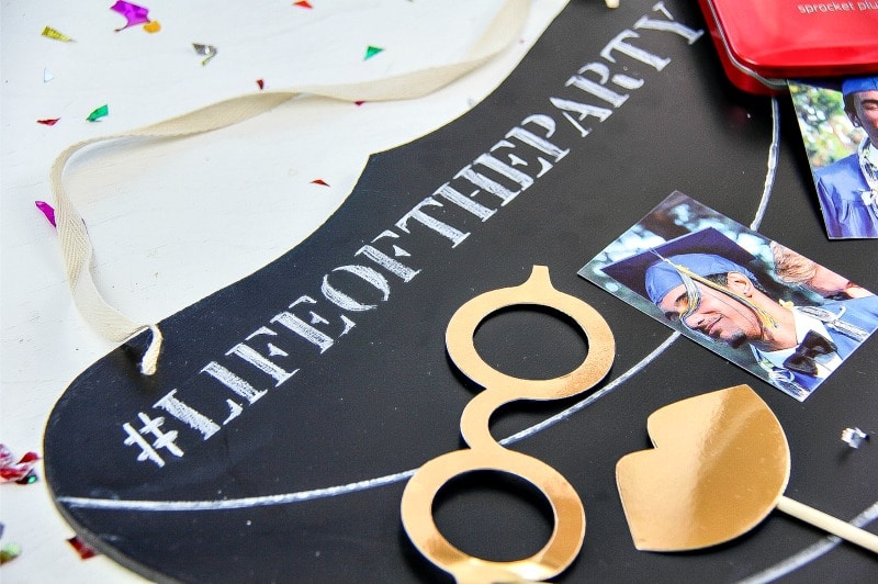 a chalkboard decoration with text reading life of the party with a couple of photos on it and paper gold glasses and lips on a stick on a white table with colored confetti on it