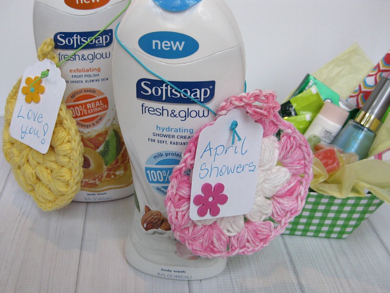 two softsoap body wash with crocheted scrubbies hanging on them with paper tags reading love you and april showers with a box of nail polish, lotion and other care items in a green and white checkered box next to them on a white wood background