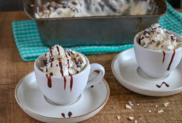almond joy ice cream in two white mugs on white saucers with more ice cream in a metal pan on a green cloth in the background, all on a brown table