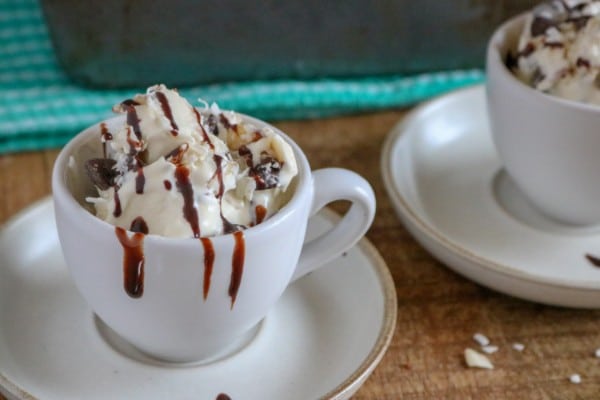 almond joy ice cream topped with chocolate sauce in white mugs on white saucers on a brown table with a metal pan of more ice cream on a green cloth in the background