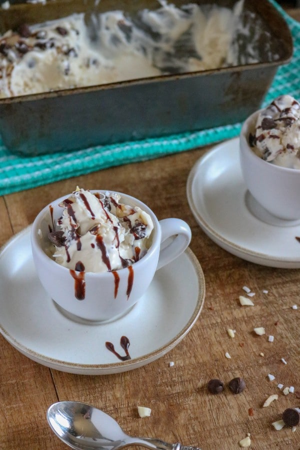 almond joy ice cream topped with chocolate sauce in white mugs on white saucers on a brown table next to a spoon with a metal pan of more ice cream on a green cloth in the background