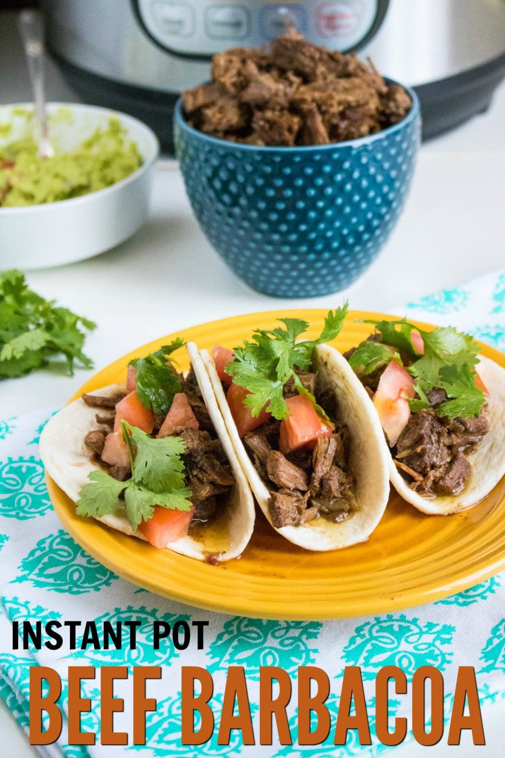 beef barbacoa in three flour tortillas topped with green garnish and chopped tomatoes on a yellow plate on a green and white cloth with a blue bowl full of bee, a white bowl of guacamole and an instant pot in the background with title text reading Instant Pot Beef Barbacoa