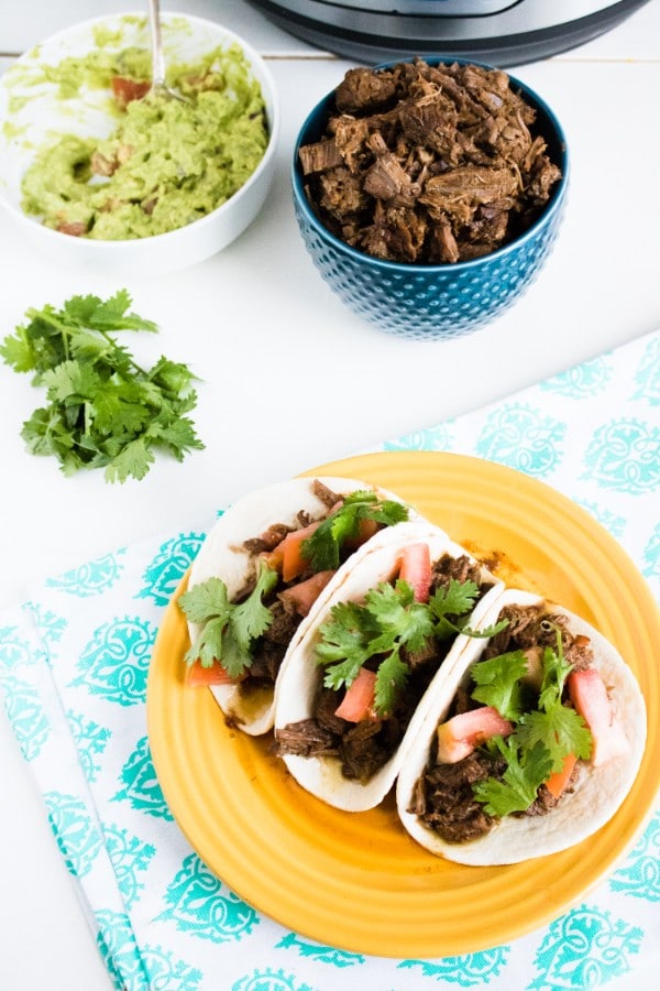 an overhead view of beef barbacoa in three flour tortillas topped with green garnish and chopped tomatoes on a yellow plate on a green and white cloth with a blue bowl full of bee, a white bowl of guacamole and an instant pot in the background