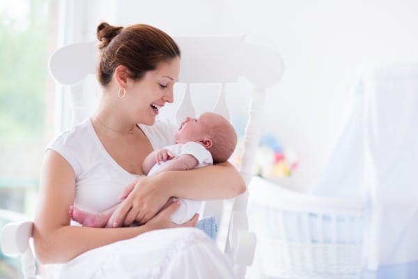 a mom smiling and looking down at the baby in her arms while they're sitting in a white rocking chair in a baby's room