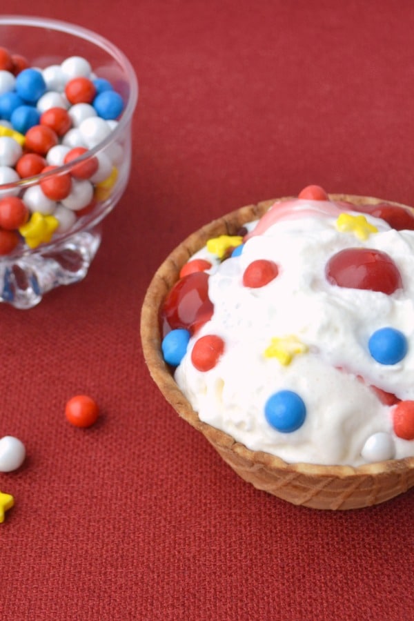 vanilla ice cream topped with red, white and blue round candies, yellow candies, and cherries in a waffle bowl next to a glass bowl of candy on a red cloth