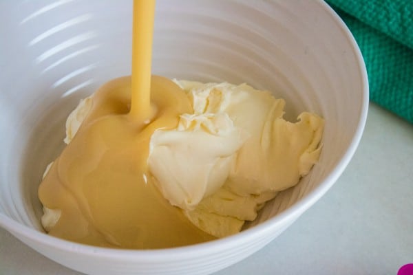 sweetened condensed milk being poured into whipped heavy cream in a white bowl on a white table next to a green cloth