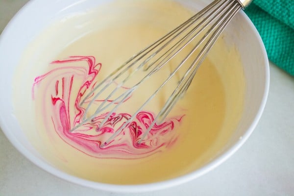 a whisk mixing in pink food coloring to the ice cream mixture in a white bowl on a white table next to a green cloth