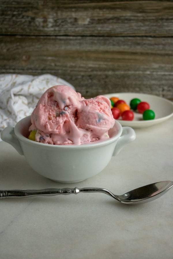 bubble gum ice cream in a white bowl next to a spoon with a cloth and a plate of bubble gum in the background on a white table