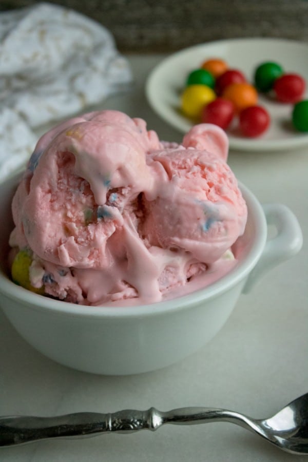 bubble gum ice cream in a white bowl next to a spoon with a cloth and a plate of bubble gum in the background on a white table