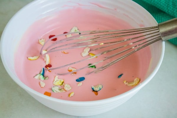 gumball pieces in the pink ice cream mixture in a white bowl with a whisk in it on a white table next to a green cloth