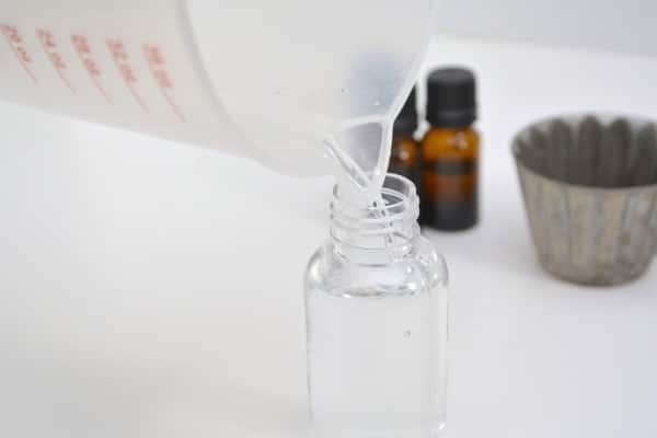 water being poured into a plastic spray bottle on a white table with bottles of essential oils and a metal cup in the background