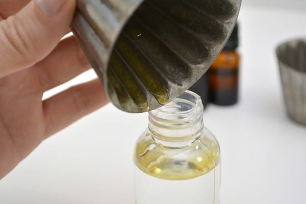 a hand pouring dish soap from a metal cup into a spray bottle on a white table with bottles of essential oils and another metal cup in the background