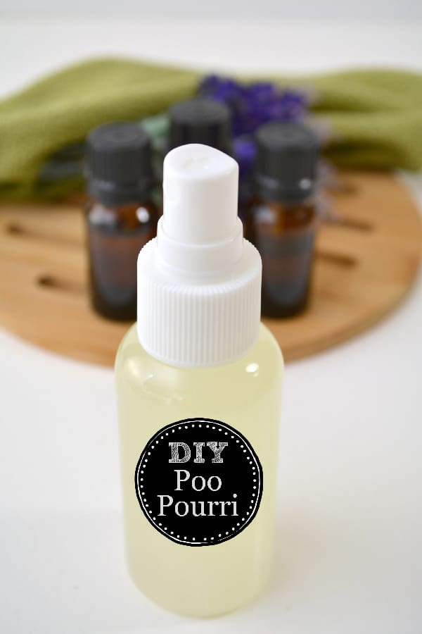 DIY Poo Pourri Spray in a plastic spray bottle on a white table with a wooden trivet, bottles of essential oils and a green cloth in the background