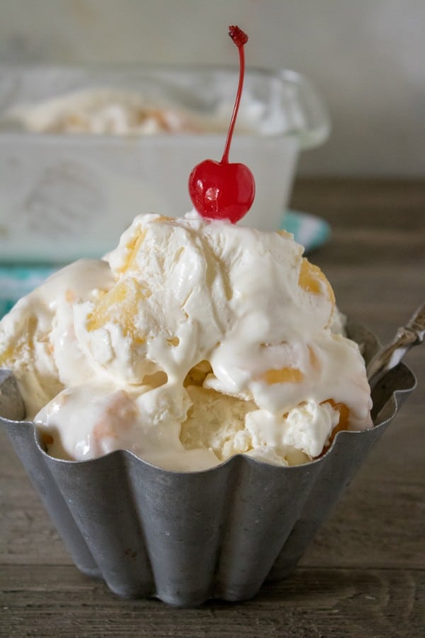 ice cream in a metal bowl topped with a cherry on a brown table with a dish of more ice cream in the background