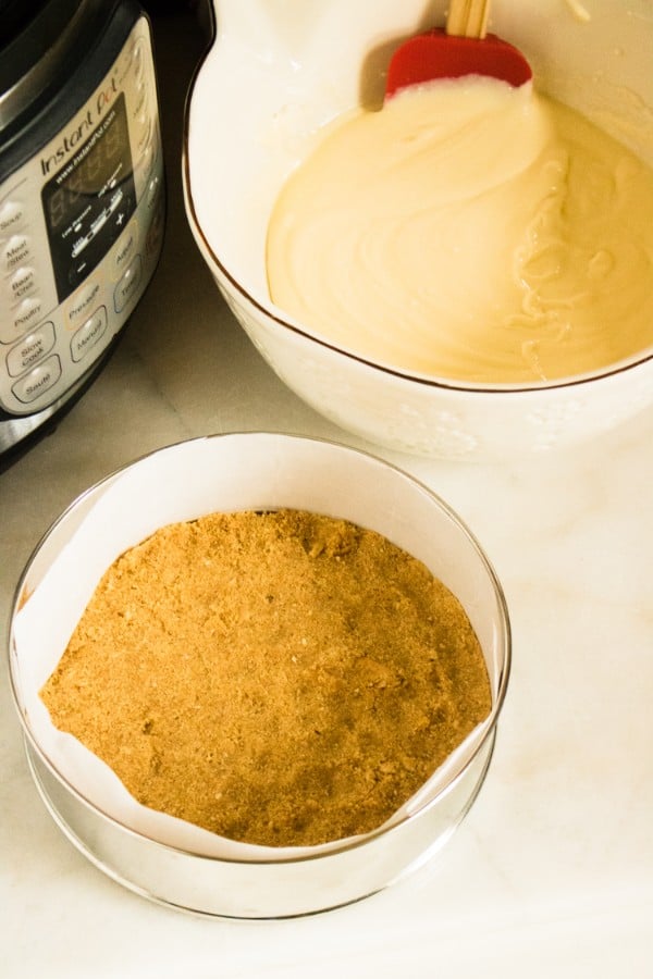 a springform pan with the cheesecake crust in it, a white bowl with peanut butter cheesecake filling and a spatula in it on a white counter next to an instant pot