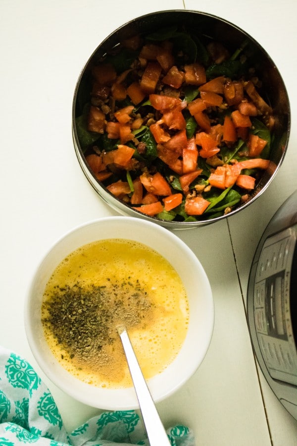 a fork in a mixture of cream, egg and seasonings in a white bowl with a pan of chopped tomatoes and spinach next to it with an instant pot in the background on a white table