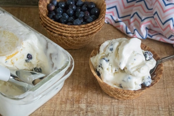 lemon blueberry ice cream in a glass dish and a waffle bowl next to a stack of waffle bowls with the top one full of blueberries on a brown table next to a cloth