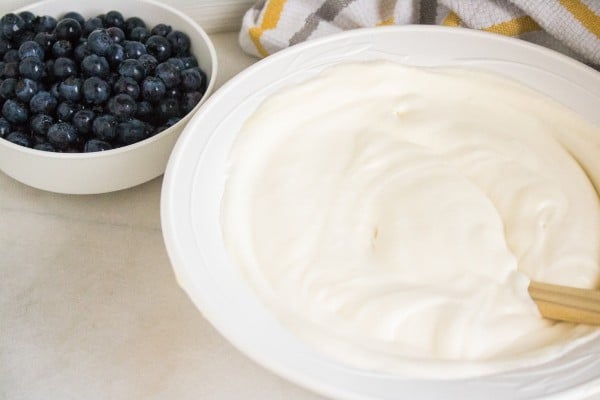 a white bowl of whipped heavy cream and condensed milk mixed together next to a white bowl of blueberries on a white counter