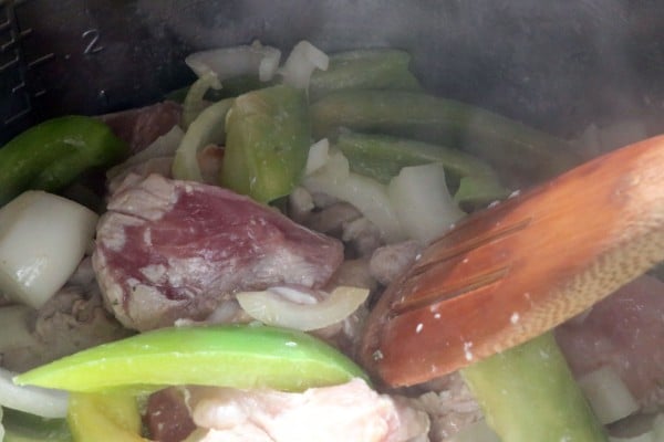 pieces of pork roast, sliced onions and green bell pepper being sauted in an instant pot with a wooden spoon