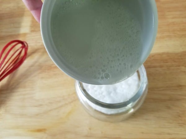 a hand pouring the diy makeup remover liquid into the jar of cosmetic pads on a wood table with a red whisk in the background