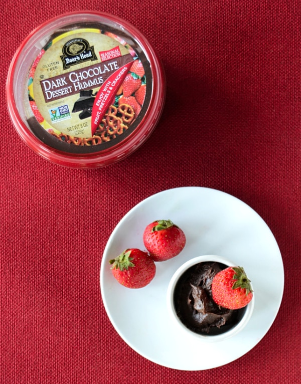 a container of dark chocolate hummus next to a white plate with strawberries and a small bowl of chocolate hummus on it, all on a red background
