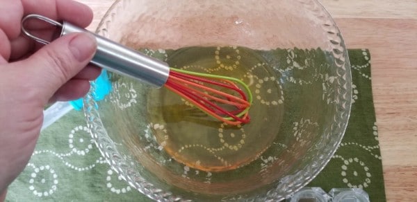 a hand using a whisk to mix the oils in a glass bowl on a green linen on a wood table