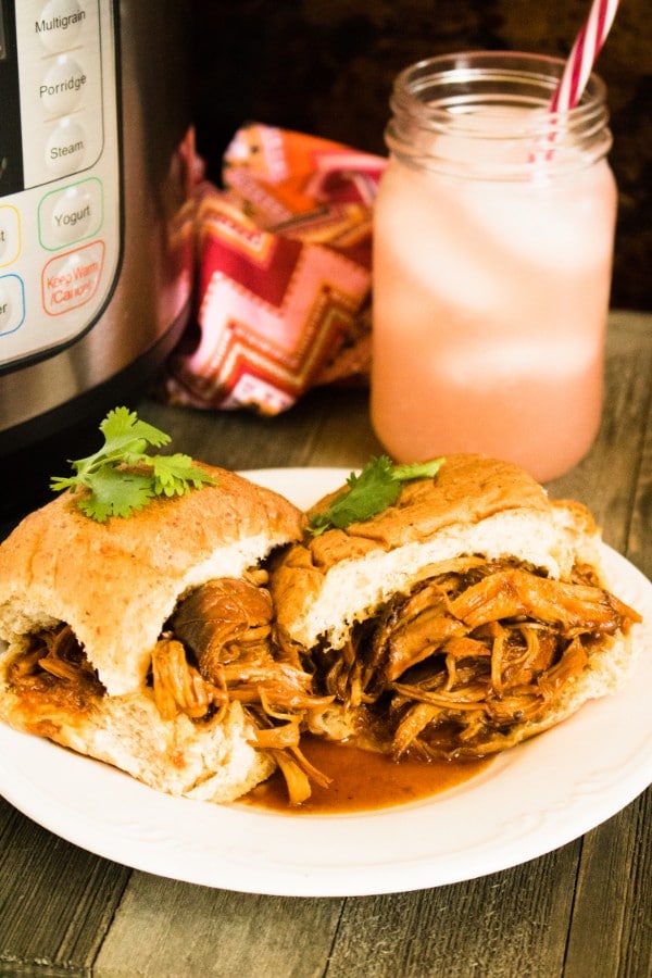 BBQ Shredded Chicken sandwiches on a white plate on a brown table with and instant pot and jar of a pink drink in the background
