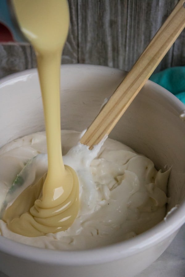 sweetened condensed milk being poured into a white bowl of whipped heavy whipping cream with a wooden spoon in the bowl