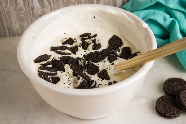 oreo pieces being folded into a white bowl of ice cream ingredients with a wooden spoon in the bowl and more oreos and a green cloth on the table