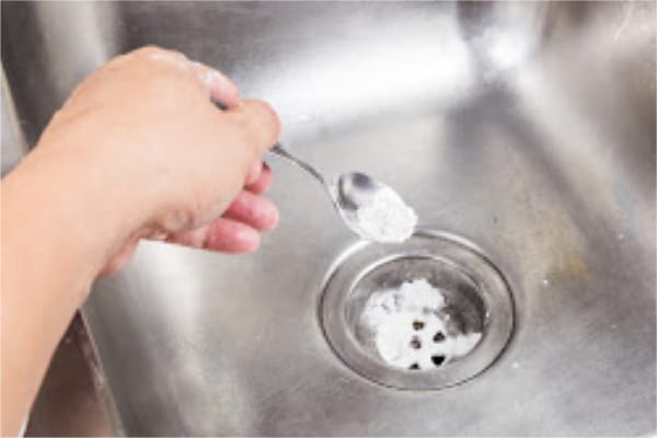 a hand using a spoon to drop baking soda down the metal kitchen sink drain