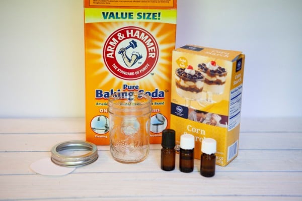 a box of baking soda and corn starch, three glass bottles of essential oils, an empty glass mason jar and lid, all on a white wood table with a white background