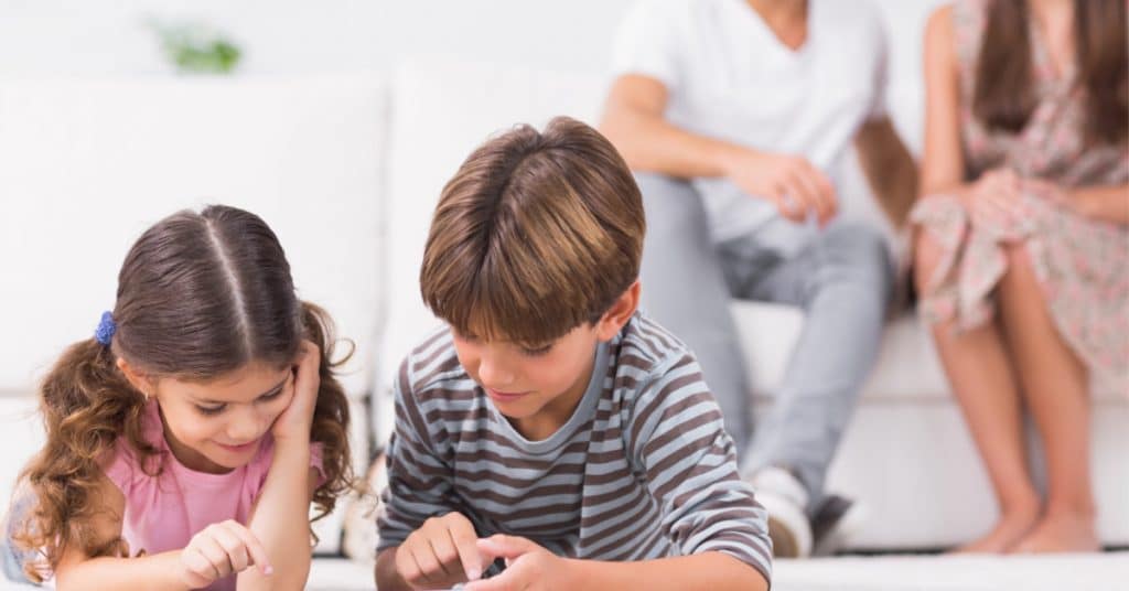 a boy and girl typing on an electronic device with their parents in the background sitting on a couch