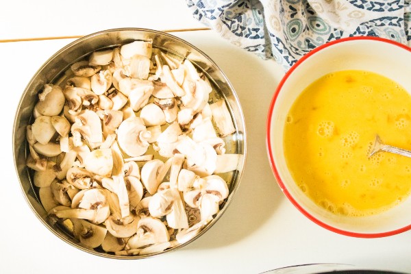 a metal bowl of sliced mushrooms next to a bowl of eggs beaten with a fork on a white table