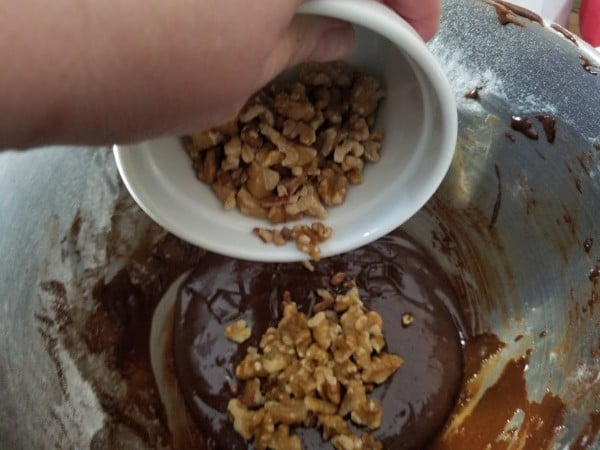a hand pouring nuts from a white bowl into the metal mixing bowl of brownie batter