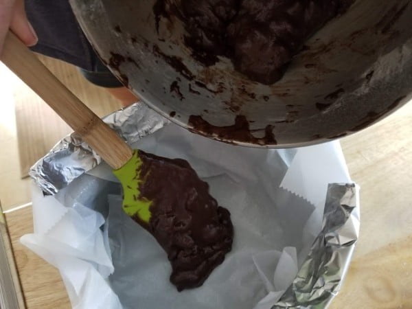 using a spatula to transfer the brownie batter from the mixing bowl to the parchment lined cake pan
