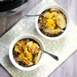 Two bowls of instant pot cheesy hamburger potato casserole topped with green onions rest on a patterned cloth, with a slow cooker in the background.
