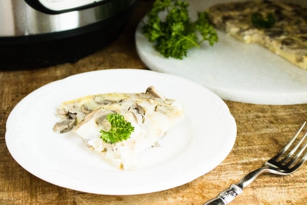 a slice of mushroom and swiss frittata on a white plate on a wood table with the rest of the frittata on a white plate in the background next to an instant pot and a fork