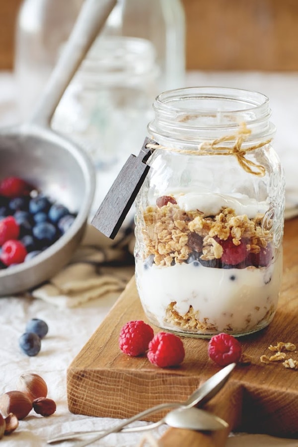 a mason jar filled with a yogurt parfait on a wooden board with more raspberries and blueberries in the background