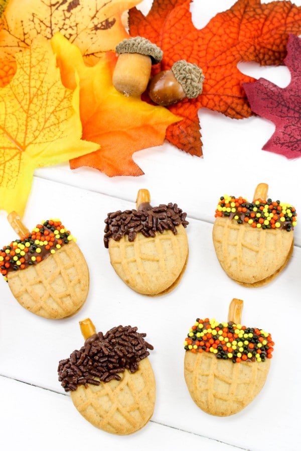halved nutter butter cookies topped with chocolate frosting, sprinkles and a broken pretzel stick to look like acorns on a white table with fake leaves and acorns in the background 