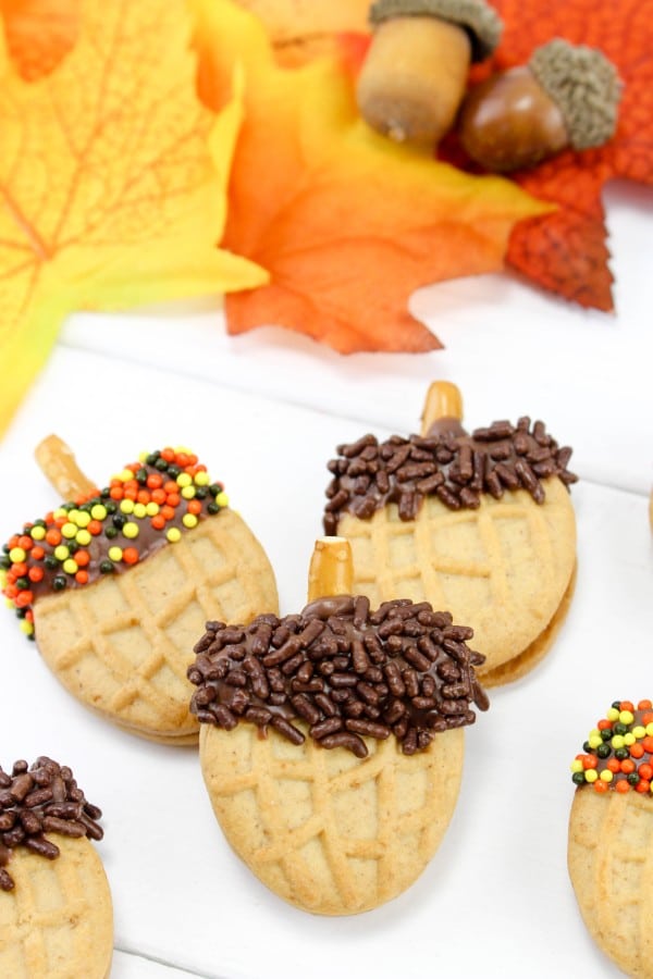 halved nutter butter cookies topped with chocolate frosting, sprinkles and a broken pretzel stick to look like acorns on a white table with fake leaves and acorns in the background 