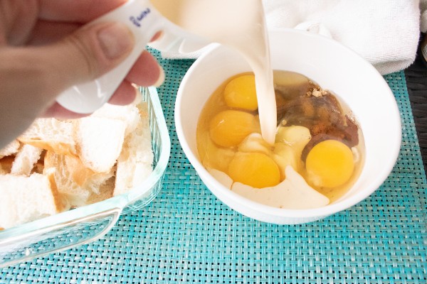 a hand pouring evaporated milk from a measuring cup into a white bowl of eggs, brown sugar and pumpkin pie spice on a blue mat next to a glass dish of torn pieces of bread