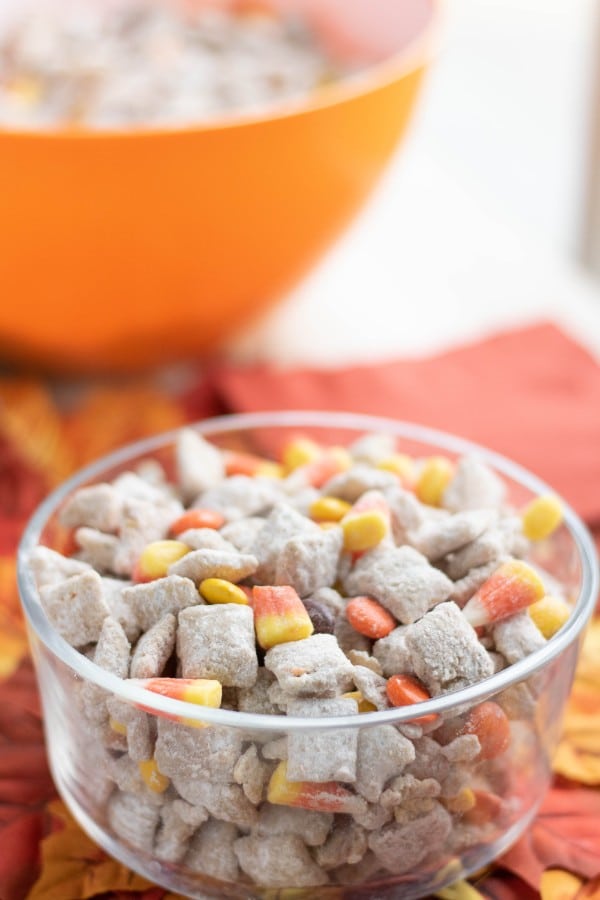 a glass bowl of chex mix with candy corn and m&Ms on fake leaves with an orange bowl of more chex mix and a napkin in the background 
