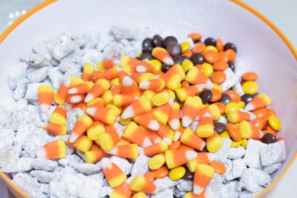 chex mix covered in powdered sugar in a white bowl topped with candy corn and m&ms